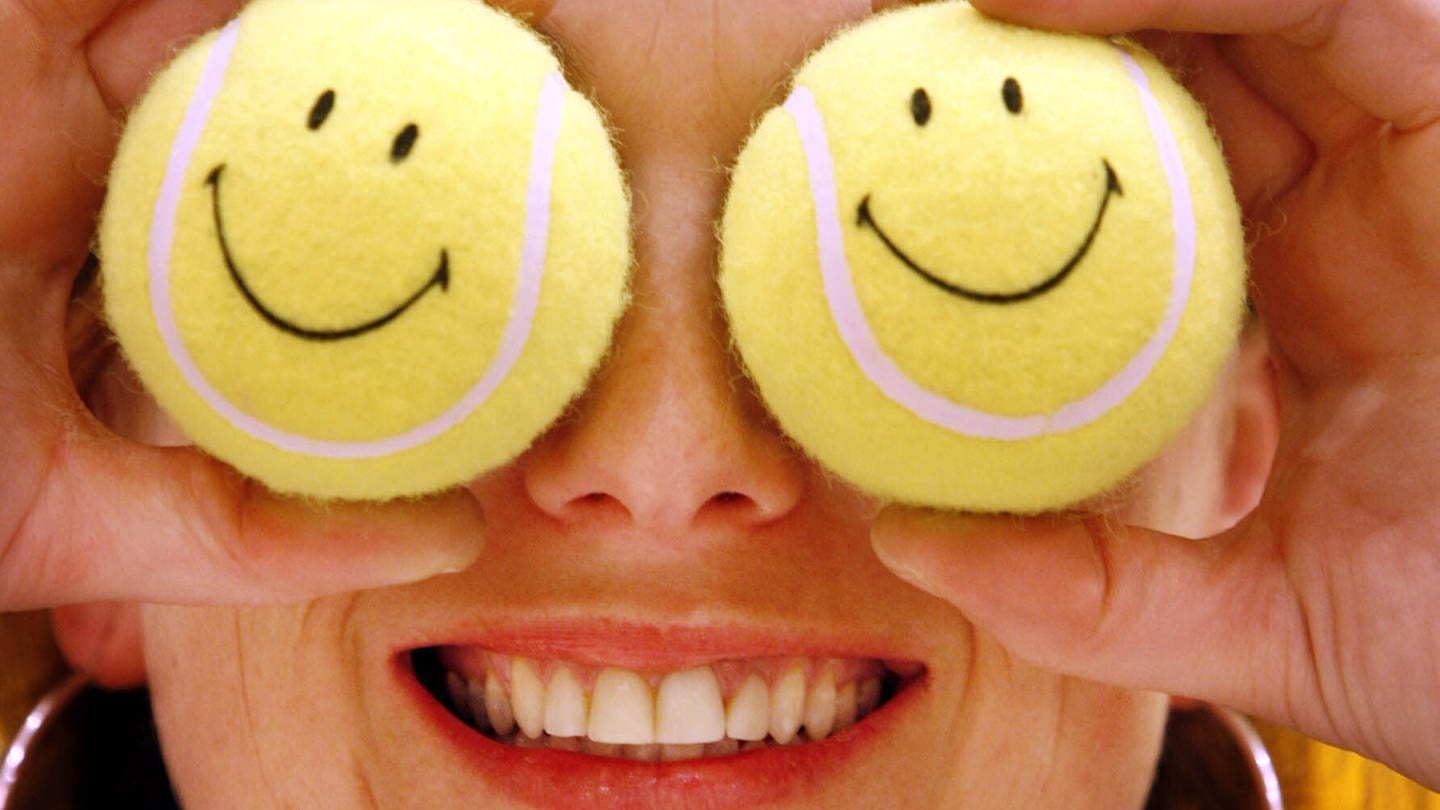 Eine Frau hält sich zwei Tennisbälle mit den aus den 70er Jahren bekannten Smileys vor die Augen.
