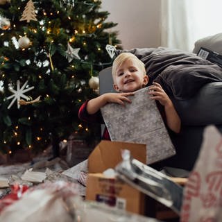 Kleiner Junge hält Geschenk hoch und lächelt vor dem Weihnachtsbaum