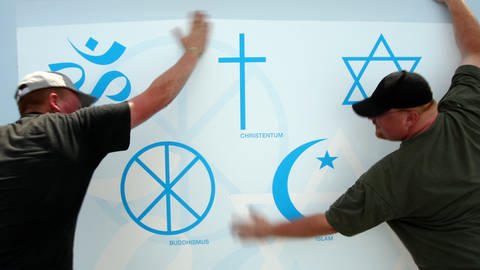 Mitarbeiter des 31. Evangelischen Kirchentages glätten ein Plakat mit fünf Symbolen von großen Weltreligionen. Auf dem Plakat sind (v.o.l.n.u.r.) die Symbole für Hinduismus, Christentum, Judentum, Buddhismus und Islam zu sehen.