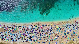 Touristen unter bunten Schirmen am türkisblauen Meer, ein Blick aus der Luft.