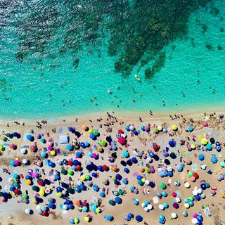 Touristen unter bunten Schirmen am türkisblauen Meer, ein Blick aus der Luft.