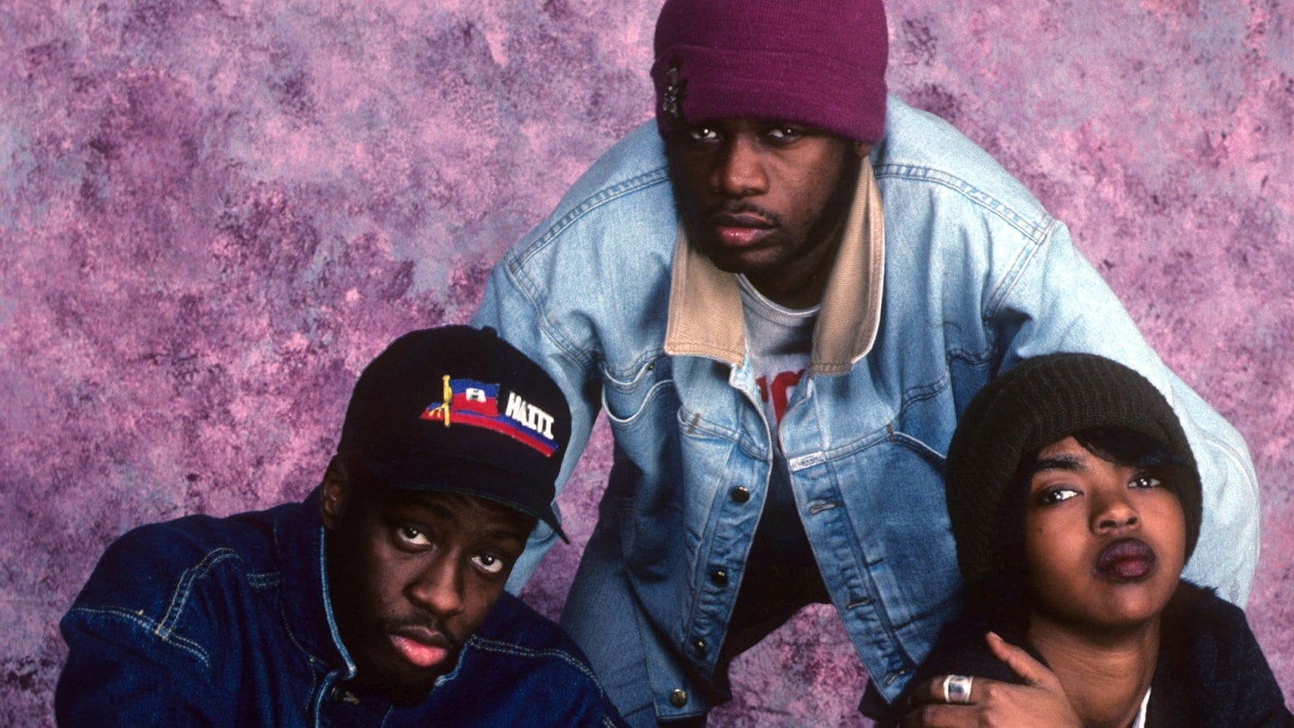 Die Band The Fugees sitzen in der Hocke auf dem Boden und schaut nach oben in die Kamera.