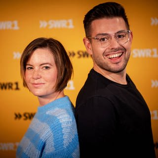 Die SWR1 Moderatoren Cora Klausnitzer und Matthias Sziedat stehen mit dem Rücken aneinander und lächeln in die Kamera.