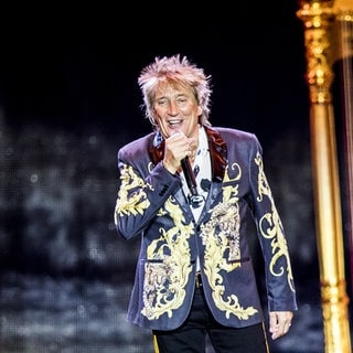 Der britische Sänger Rod Stewart trägt einen dunkelblauen Anzug mit goldenem Muster. Er singt in ein schwarzes, kabelloses Mikrofon.