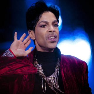 Der Sänger Prince steht auf einer Bühne. Er trägt ein rotes Samtoberteil mit glitzernden Ketten.