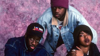 Die Band The Fugees sitzen in der Hocke auf dem Boden und schaut nach oben in die Kamera.