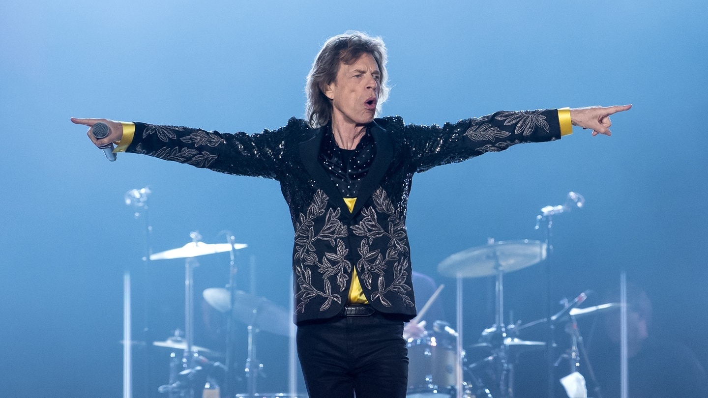 Mick Jagger steht auf einer Bühne mit weit ausgebreiteten Armen und dirigiert das Publikum.