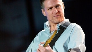 Brian Adams steht auf einer Bühne und spielt Gitarre.