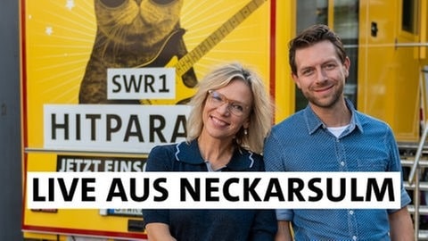 Stefanie Anhalt und Corvin Tondera-Klein stehen vor dem gelben SWR1 Bus und lächeln in die Kamera. Sie senden für die SWR1 Hitparade 2024 aus Neckarsulm.