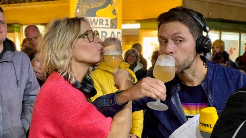 SWR1 Moderator:innen Stefanie Anhalt und Corvin Tondera-Klein trinken mit überkreuzten Armen aus Gläsern.