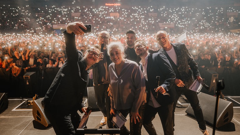 Die SWR1 Moderator:innen machen ein Selfie auf der Bühne mit der vollen Halle im Hintergrund.