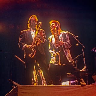 Zwei Musiker stehen im goldenen Scheinwerferlicht auf der Bühne und spielen Saxophon - Was wären die größten Hits aller Zeiten ohne ein richtig gutes Saxophon-Solo? SWR1 Baden-Württemberg hat für euch die bekanntesten Saxophon-Songs zusammengestellt: Von "Year of the Cat" von Al Stewart über "Baker Street" von Gerry Rafferty, "Careless Whisper" von George Michael und "Private Dancer" von Tina Turner bis hin zu einer überraschenden Version von "Take Five".