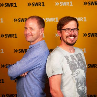 Die SWR1 Moderatoren Patrick Neelmeier und Ingo Lege stehen mit dem Rücken aneinandergelehnt und lächeln in die Kamera.