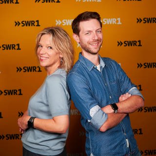 Die SWR1 Moderatoren Stefanie Anhalt und Corvin Tondera-Klein stehen mit dem Rücken aneinandergelehnt und lächeln in die Kamera.