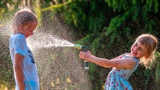 Abkühlung im Sommer: ein Mädchen bespritzt einen Jungen mit Wasser aus dem Gartenschlauch, beide lachen und freuen sich