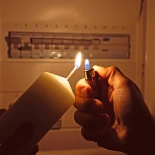 Stromausfall: ein Mann zündet mit einem Feuerzeug eine Kerze an, dahinter sieht man einen Sicherungskasten - die Sicherungen sind offensichtlich alle in "An"-Stellung