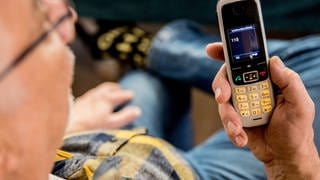 Enkeltrick oder falscher Polizist: Ein Mann sitzt in seiner Wohnung und hält ein Mobiltelefon in der Hand auf dem Display des Telefons ist die 110 zu lesen.