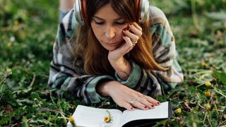 Eine Frau liegt lesend auf einer Wiese. Ein gutes Buch lesen macht den Urlaub perfekt. SWR1 verrät, welche Bücher an Pfingsten oder bei der Reise im Sommer 2023 mit dabei sein müssen.