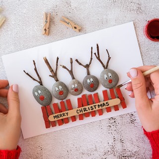 Wundervolles Winterwissen - der SWR1 Adventskalender: Warum macht uns basteln im Advent und an Weihnachten glücklich? Das Bild zeigt zwei Frauenhände, die Wäscheklammern auf einer Weihnachtskarte mit roter Farbe bemalt. Sie sind zusammen mit kleinen Steinen und Zweigen auf der Karte aufgeklebt - es sieht so aus, als schauten Rentiere über einen Zaun. Ein Schriftzug sagt "Merry Christmas"