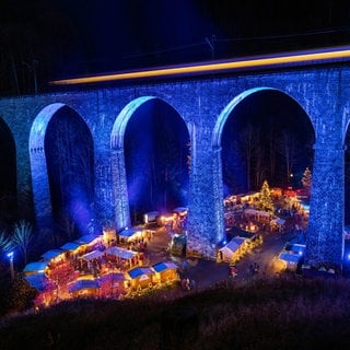 Wundervolles Winterwissen - der SWR1 Adventskalender: Woher kommt der Weihnachtsmarkt. Das Bild zeigt den Weihnachtsmarkt in der Ravennaschlucht. Die Höllentalbahn fährt über das blau beleuchtete Viadukt über der Ravennaschlucht, während darunter der Weihnachtsmarkt stattfindet.