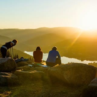 Wandern in Baden-Württemberg - Baden-Württemberg hat tolle Wanderziele: Schwarzwald, Schwäbische Alb, rund um den Bodensee. Diese Tipps sind perfekt für eine Wanderung im Urlaub oder den Ausflug am Wochenende. Das Bild zeigt Wanderer, die auf Felsen sitzen und im Schwarzwald über einen See auf das Panorama im Sonnenuntergang schauen.