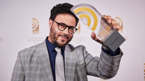 SWR1 Baden-Württemberg Moderator Nabil Atassi hält seinen Preis in der Hand. Die Folge "Pflege am Limit, Krankenschwester auf einer Corona-Station" des Podcasts "Corona Helfer:innen" wurde mit dem Deutschen Radiopreis ausgezeichnet.
