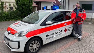 Daniel Müller steht neben dem Einsatzwagen von "Helfer vor Ort" - er hält einen Defibrillator in der Hand.