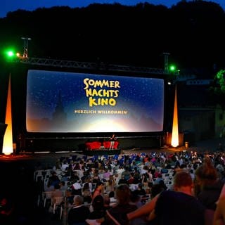 Menschen sitzen in Tübingen in einem Open-Air-Kino vor einer großen Leinwand, auf der "Sommernachtskino, Herzlich Willkommen" steht. Bei SWR1 Baden-Württemberg bekommt ihr eine Übersicht über das Programm der Open Air Kinos in BW - vom Filmklassiker bis zum aktuellen Hollywood Blockbuster. Egal ob in Heilbronn, am Bodensee, in Freiburg, Stuttgart, Esslingen, Ulm oder Bad Mergentheim: Freiluft Kino an einem lauen Sommerabend ist ein ganz besonderes Erlebnis!