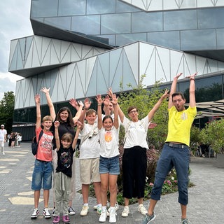 Der perfekte Tag mit SWR1 bei der Experimenta in Heilbronn. Die Gewinner-Gruppe und SWR1 Moderator Corvin Tondera-Klein stehen vor dem glasverkleideten, futuristisch aussehenden Gebäude und recken die Arme in die Höhe.