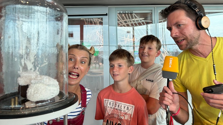 Der perfekte Tag mit SWR1 in der Experimenta in Heilbronn: Experimenta-Edutainerin Kelly, Henry, Erik und SWR1 Moderator Corvin Tondera-Klein schauen auf eine große Glasglocke. In ihr steht ein explodierter Schokokuss, weil in der Glasglocke ein Vakuum herrscht.