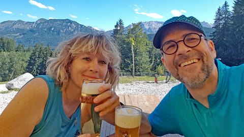 Kris und Frank beim Wandern - sie prosten sich mit Biergläsern zu, im Hintergrund die Allgäuer Alpen. SWR1 Neuanfang: Kris rettet mit einer Organspende ihrem Mann Frank das Leben.