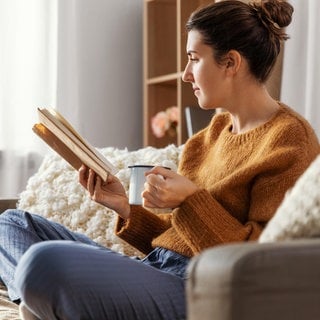 Eine Frau sitzt im Schlafanzug auf einer Couch und liest ein Buch. In der Hand hält sie eine Tasse Kaffee.