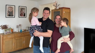 Familie Thiessen im Wohnzimmer: Papa Jakob mit Tochter Elisa auf dem Arm, Mama Melanie rechts neben ihm hat Sohn Jonas auf dem Arm.