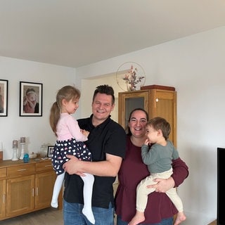 Familie Thiessen im Wohnzimmer: Papa Jakob mit Tochter Elisa auf dem Arm, Mama Melanie rechts neben ihm hat Sohn Jonas auf dem Arm.