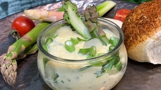 Auf einem Tisch steht ein rundes Glas mit Spargel-Käse-Aufstrich, Spargel und einem Brötchen.