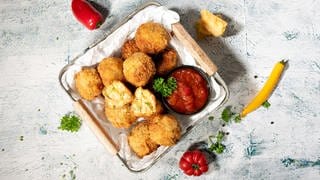 Auf einem Tisch steht ein Glasbehälter mit einer Portion Käsespätzle-Arancini. Daneben ein Schälchen mit einer Salsa-Sauce.