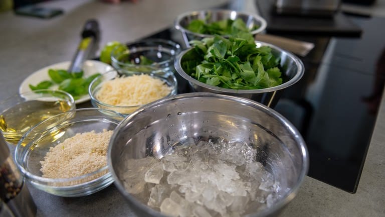 Alle Zutaten für ein grünes Kräuterpesto stehen in Schüsseln auf einer Arbeitsplatte in einer Küche.