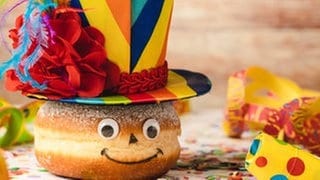 SWR1 Pfännle meets Fasnacht - wir feiern Fasnet, Fastnacht, Fasching und Karneval kulinarisch! Küchenkäpsele und Rezepte für Fasnachtsküchle, Nonnenfürzle und mehr.  Das Bild zeigt einen Berliner  Krapfen im Faschingskostüm: ein bunter Hut mit Federn, Konfetti und Luftschlangen