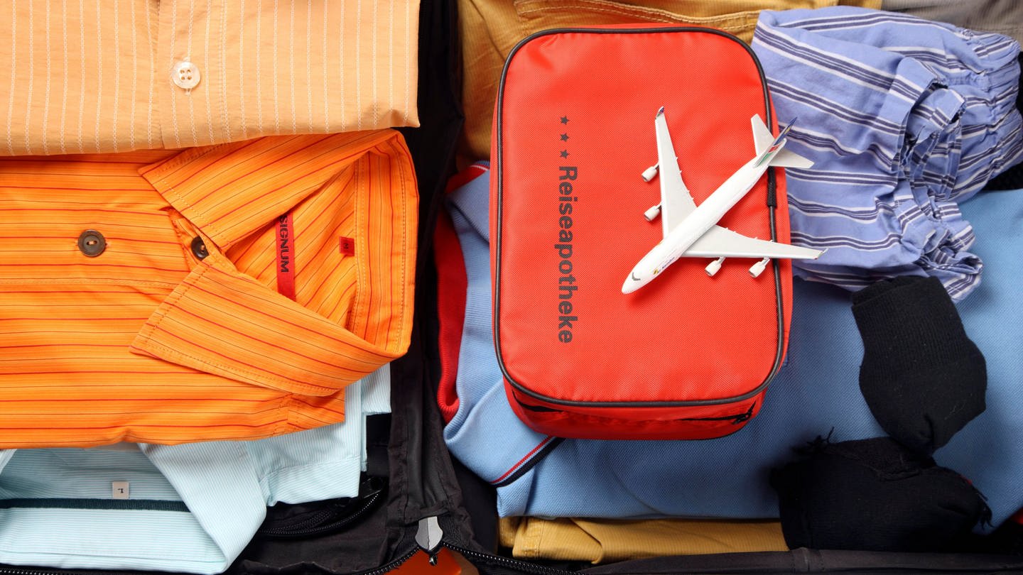 Eine Tasche mit der Aufschrift Reiseapothke liegt in einem gepackten Koffer. Auf ihr befindet sich ein kleines Flugzeug