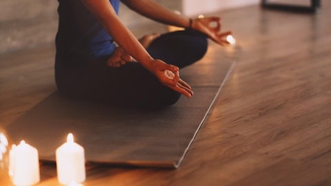 Eine Frau macht auf einer Matte eine Yoga übung im Schneidersitz. Neben ihr leuchten Kerzen