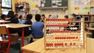 Ein Rechenschieber steht auf einem Tisch im Klassenzimmer in einer Grundschule. Im Hintergrund sind die Tafel und Kinder zu erkennen.