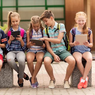 Fünf Schulkinder sitzen auf einer Bank und schauen auf ihre Tablets