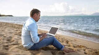 Mann sitzt mit einem Laptop am Strand