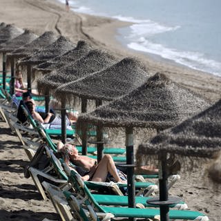 Menschen liegen am Strand von Spanien auf Strandliegen mit Sonnenschirmen aus Stroh