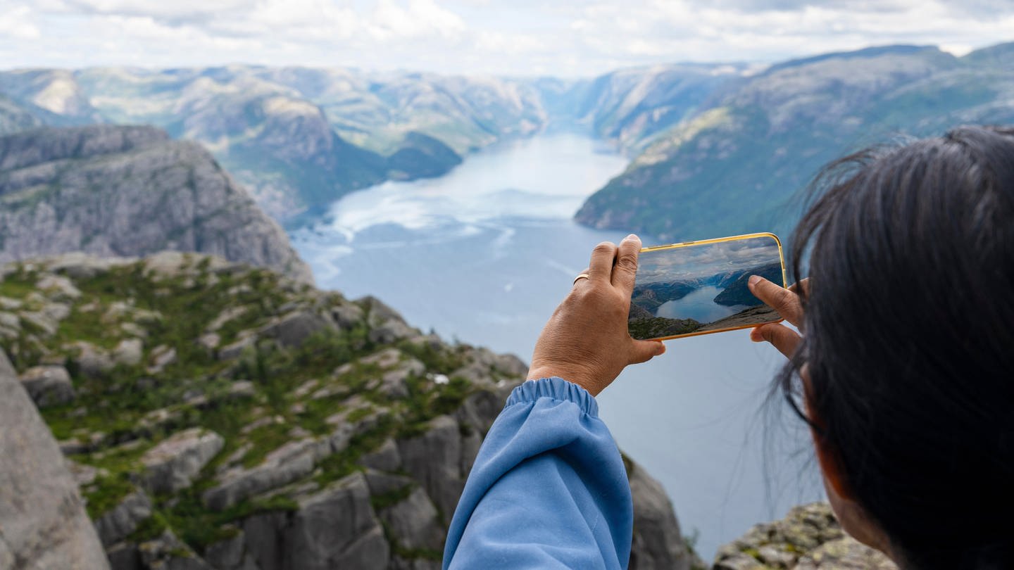 Frau steht auf dem Berg mit dem Felsen Preikestolen mit aussicht auf den Fjord Lysefjord und macht ein Foto mit dem Smartphone.