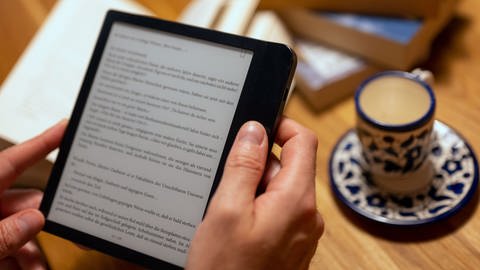 Eine Frau list mit dem Tolino ein E-Book | E-Book-Reader: Was können Tolino und Kindle?