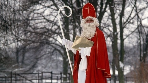 Ein Mann mit Nikolauskostüm, Hirtenstab und Bischofsmütze in einer verschneiten Landschaft in Deutschland. | So kommt der Wunschzettel bei Christkind und Weihnachtsmann an