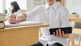 Ein Kind sitzt an einem Tisch in der Schule und schaut auf sein Smartphone | Sollte es ein Smartphone-Verbot an Schulen geben?