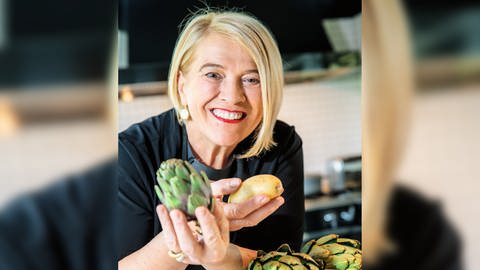 Spitzenköchin Léa Linster hält Gemüse in der Hand und lacht in die Kamera. 