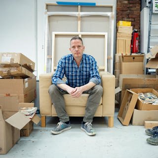 Der Maler Norbert Bisky sitzt auf einem Sofa in seinem Atelier. Norbert Bisky ist einer der wichtigsten zeitgenössischen deutschen Künstler, seine Werke sind in den bedeutendsten Museen der Welt zu sehen etwa dem MOMA in New York. Er verbindet Gemälde- und Bühnenkunst.